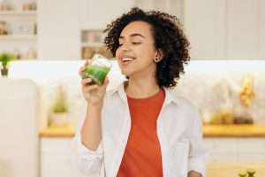 Happy woman with curly hair dressed in red and white, standing in the kitchen drinking healthy green juice